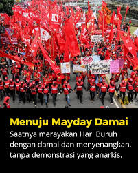 A large crowd of marchers waving red flags in a May Day rally; banner and caption promote a peaceful Labour Day without anarchic demonstrations.