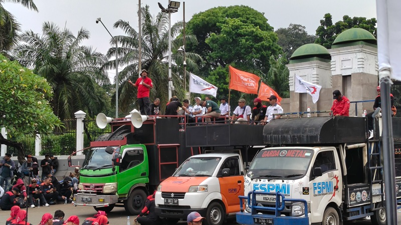 Group of people on flatbed trucks carrying flags, part of a street parade or protest, with onlookers seated nearby and palm trees in the background.