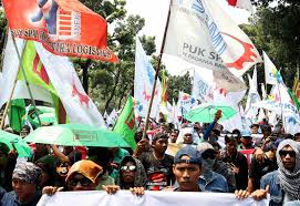 Large crowd of protesters marching with colorful flags and banners at a street demonstration under a sunny sky.