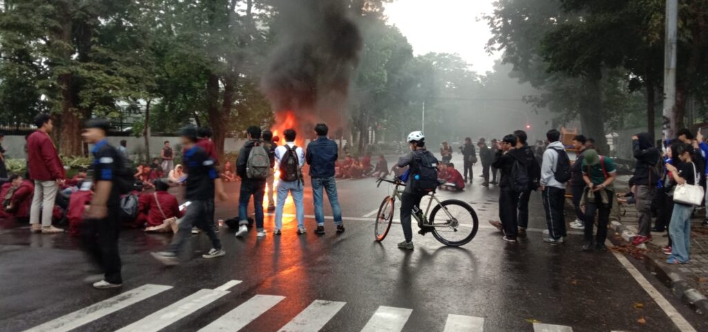 Crowd of bystanders on a wet street around a large fire with dark smoke during a protest or confrontation.